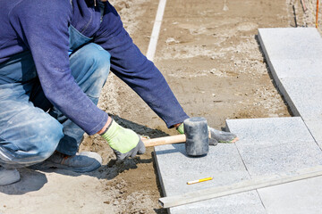 A worker places granite blocks on the sidewalk with a rubber hammer on a marked area.