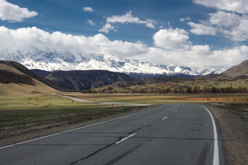 Mountain steppe valley road path with spring green grass and ranges of  snow mountains rocks on a horizon skyline under blue sky