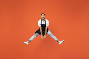 Fototapeta na wymiar Full length young man barista bartender barman employee in black apron white tshirt work in coffee shop jump high with outstretched hands isolated on orange background. Small business startup concept.