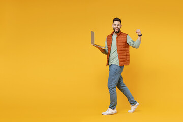 Young freelancer happy caucasian man in orange vest mint sweatshirt glasses using laptop pc computer chat online browsing internet do winner gesture clench fist isolated on yellow background studio.