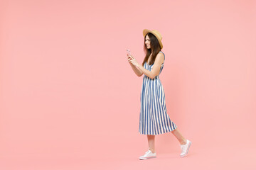 Full length side view young fun caucasian woman in summer clothes striped dress straw hat using mobile cell phone chat online internet walk isolated on pastel pink color background studio portrait