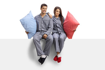 Young man and woman in matching pajamas sitting on a white panel and holding pillows