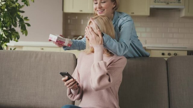 Smiling young adult daughter makes an unexpected surprise to her middle aged attractive mom, gives her a gift, covering her eyes with self hand, sitting at living room. Friendship of mom and daughter