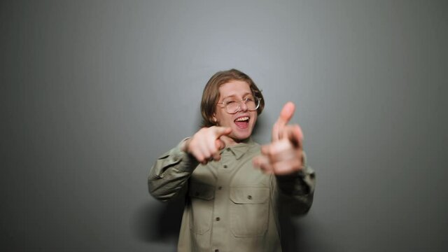 Joyful young man nerd in a shirt and glasses dances on a background of a gray wall and shows his fingers to the camera with joy.