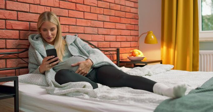 Young pregnant woman relaxing on bed using smartphone texting message to husband. Beautiful elegant lady motherhood with bare belly laying on bed sharing ultrasound scan picture on mobile phone.