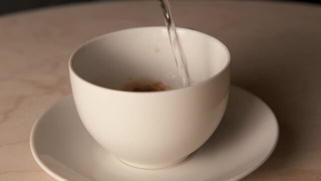Boiling water is poured into a white cup of instant coffee on a white saucer on a wooden table. The camera rotates around the table 360 degrees