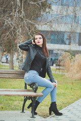 Vertical shot of a young brunette female in a leather coat posing sitting on a bench