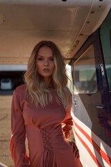 Pretty blond woman with wavy hair wearing pink silk classy dress posing near a private plane