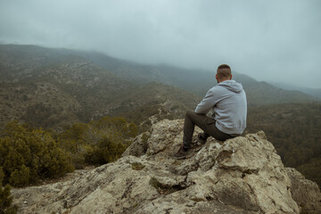 Man from behind in a gray jacket sitting on a rock on top of a mountain in autumn with mist