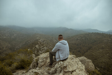 Man from behind in a gray jacket sitting on a rock on top of a mountain in autumn with mist