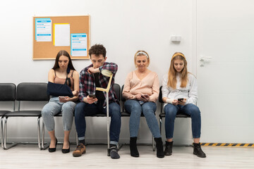 Patients sit in a clinic waiting for their turn to go to the doctor's office. They look at their cell phone while waiting.