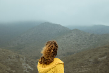 Woman from behind with a yellow jacket sits on a rock on top of a mountain in autumn with mist