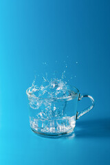 Clear cold water in a glass cup on blue background. Splash from falling ice