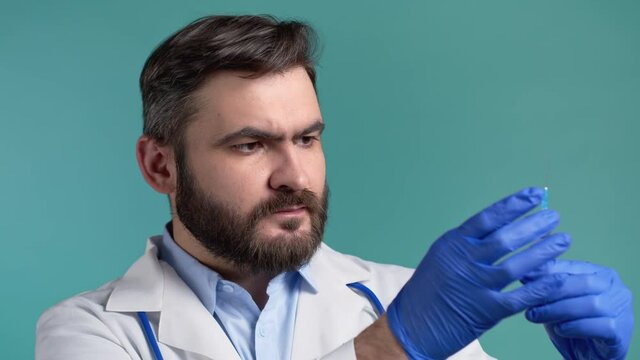 Doctor in gloves flicks syringe prior to drug injection, to collect and release the air out of syringe. Bearded man on blue studio background. Healthcare, vaccination, medicine concept.