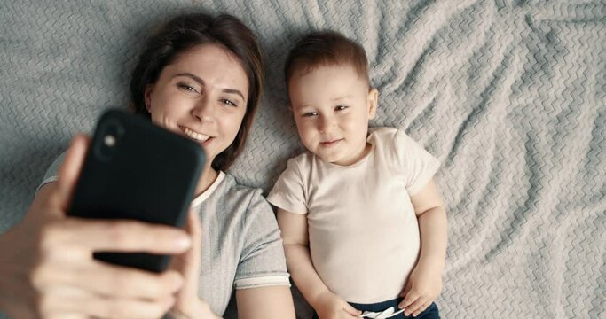 Mother and her baby boy making a selfie or video call to father or relatives in a bed. Concept of technology, new generation, family, connection, parenthood. Top view, slow motion.