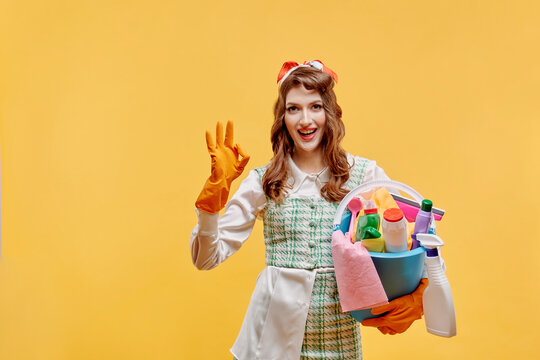 The happy cleaning lady shows the ok gesture and looks at the camera. A bottle of household chemicals and cleaning products. A sponge for washing dishes and a rag. Pin-up style. Yellow background.