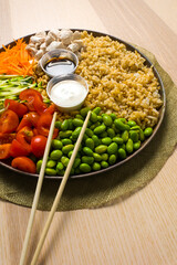detail with chopsticks of a dish with Asian cuisine ingredients: brown rice, chicken, carrots, cherry tomatoes, zucchini, edamame, yogurt sauce, soy sauce