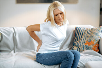 Upset frustrated mature woman suffering from backache, unhappy senior blonde sitting on a sofa at living room, feeling discomfort because of pain in back, have joint problems, need rest. - obrazy, fototapety, plakaty