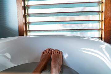 Bathtub feet soaking in warm water woman taking a warm bath at luxury bathroom villa. Window view.