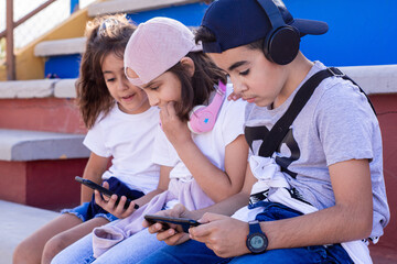 Three Caucasian children sit together on bleachers, laughing as they look at their smartphones.