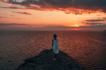 a little girl in a white dress stands on a cliff above the sea at a beautiful sunset back view