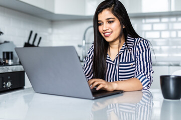 happy young indian woman using laptop in the kitchen