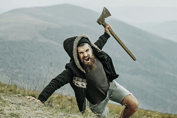 Brutal man lumberjack hipster with long beard and moustache in coat and hood with axe in hand on...