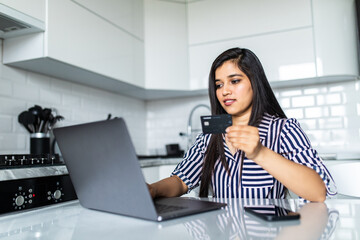Smiling indian woman online shopping using laptop and credit card in kitchen