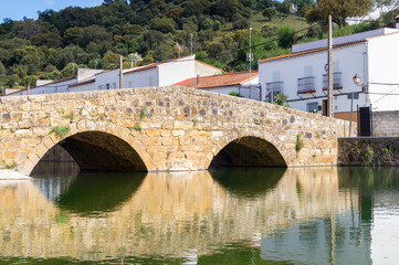 Roman stone bridge over the Galindon river in San Nicolas del Puerto (Seville, Spain). Nice old bridge for the pedestrian crossing in a rural village with white houses and nature in the background.