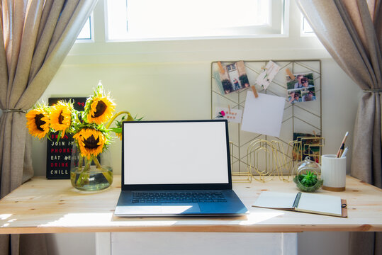 Home office workspace. Wooden desk with laptop mockup white empty screen, mood board with pined notes and photos, sunflower bouquet in the vase. Sunny day. Work from home workspace. Copy space.