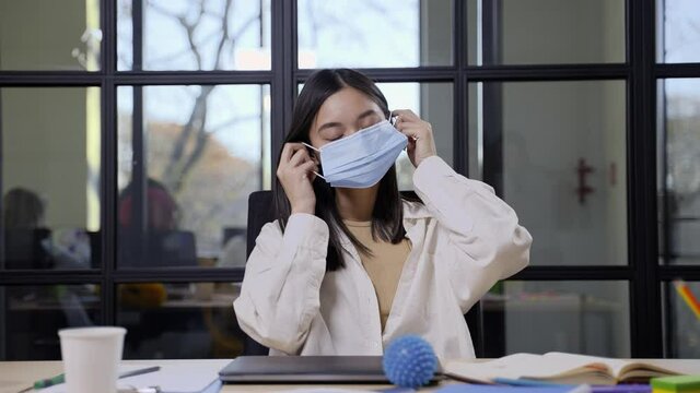 Close-up of attractive korean female wearing medical mask posing on camera at office table during covid-19 epidemic. Masked freelancer sitting at workstation in modern office with glass partitions