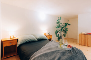 Ordinary bedroom in loft apartment in wooden style with double bed, bedside tables and two glasses of wine with bottle on tray.  Bedroom is lit by bright light mounted on wall and two bedside lamps.