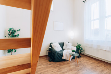 Bright loft apartment with large window, wooden floor and built-in staircase. The room has decorative plants, a sofa, lamps and couple of fluffy pillows.