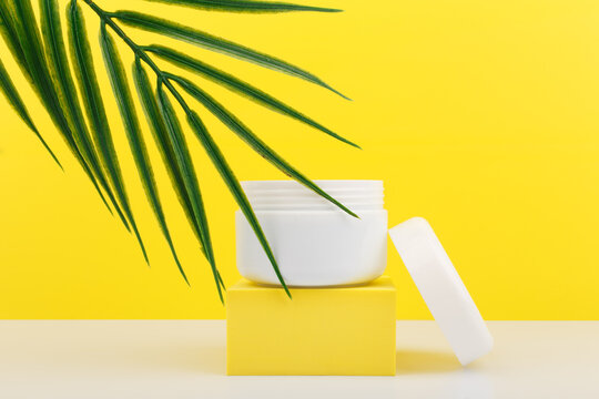 White Opened Cosmetic Jar On Yellow Podium With Palm Leaf Against Bright Yellow Background. Cosmetic Jar For Face Mask, Cream, Scrub Or Balm