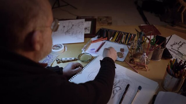 View from behind of an artist working in the studio in a creative environment. The older man is drawing a portrait with watercolors using a brush.