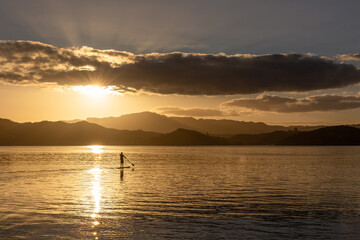 A photo of a silhouette of a person sailing on paddleboard on the calm surface of the sea during the golden hour of sunset