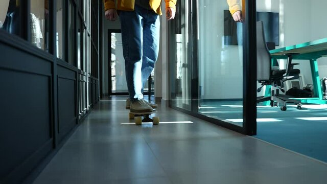 Active worker in casual clothing riding board down corridor of contemporary office with glass partitions. Sporty hipster skating around office coworking space, picking board up and greeting colleagues