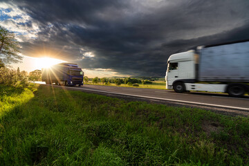 Motion blurred white truck and blue truck driving on the asphalt road in rural landscape in the rays of the sunset with dark storm cloud