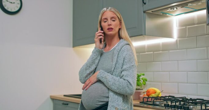 Young pregnant woman talking to her husband using phone at home. The pregnant girl communicates her family via cell phone information. A caring mother strokes a pregnant tummy hand.