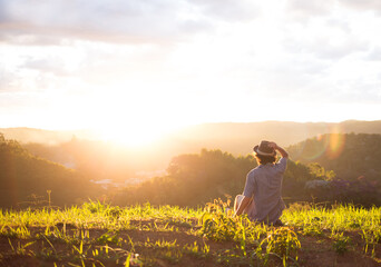 Fototapeta premium Traveler wearing hat sitting on the top of a mountain with the sunset in the background.