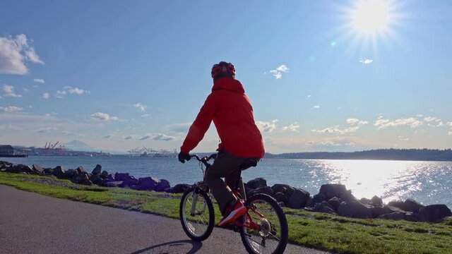 Senior man riding bike on Seattle waterfront trail with bright sun behind, healthy lifestyle.