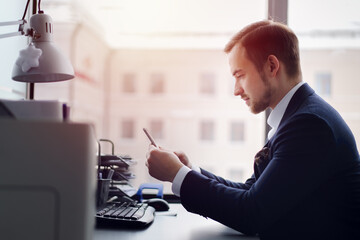 The businessman at the table in the office looks at the phone. On the table are a printer, a lamp, a paper segregator, the keyboard.