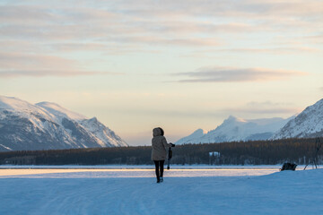 Woman holding a tripod and camera walking along a frozen lake in northern Canada on a cold, sunset afternoon in spring time April. Wearing beige jacket and black pants. 