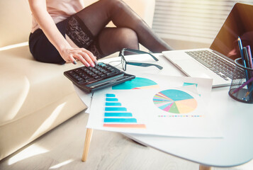 Businesswoman in black stockings and high heels sits on the couch near the table with a laptop and financial charts