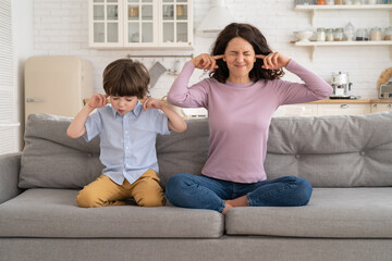Frowning mom and son sit on couch with closed eyes and cover ears from noisy music or fight sounds...