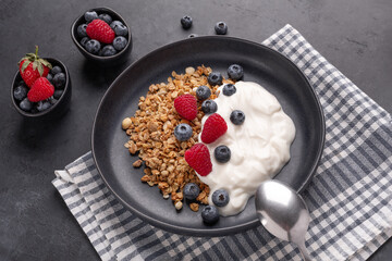 Yogurt with baked granola and berries in black ceramic plate on dark stone background. Healthy breakfast - Image