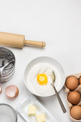 Brown eggs in carton container. Butter and whisk on plate. Egg yolk with flour in bowl.