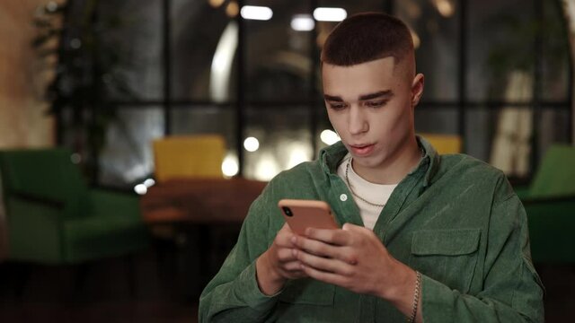 Crop view of stylish man looking amazed and saying wow while using smartphone in cafe. Handsome guy with chainlet scrolling and browsing internet while showing emotions.