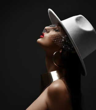 Profile portrait of rich gorgeous woman in wide-brimmed white hat with veiling, massive metal collar and earrings holding head up with eyes closed over black background