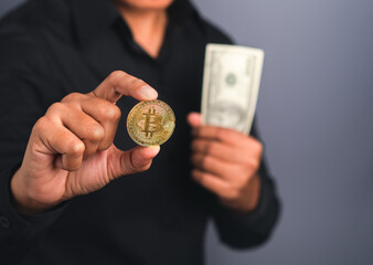 Portrait of an investor guy showing a bitcoin golden coin and US banknote with a gray background while standing in the studio. Space for text. Virtual cryptocurrency and future money concept
